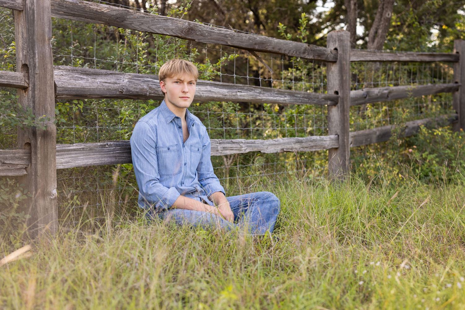 2027 senior boy in a denim shirt and jeans sitting cross-legged in the grass beside a rustic wooden fence outdoors.