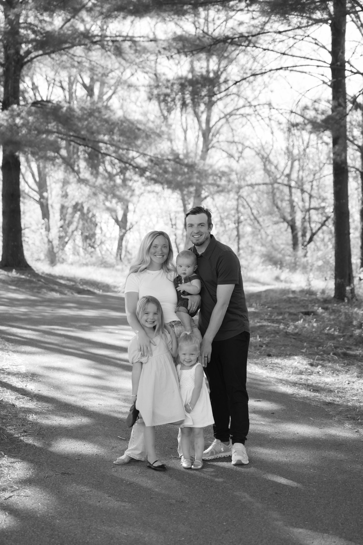 black and white family photo of parents and three toddlers smiling at the camera with a wooded backdrop in hudson wi