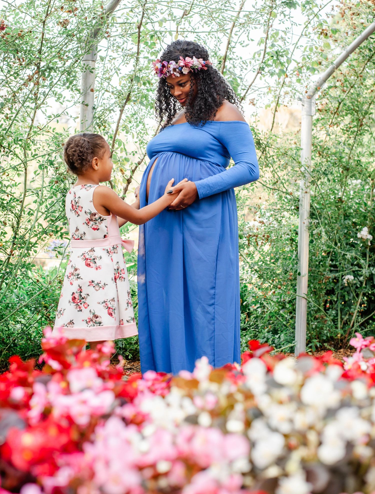 Pregnant woman in blue gown with flower crown poses with partner and young daughter in lush garden setting.