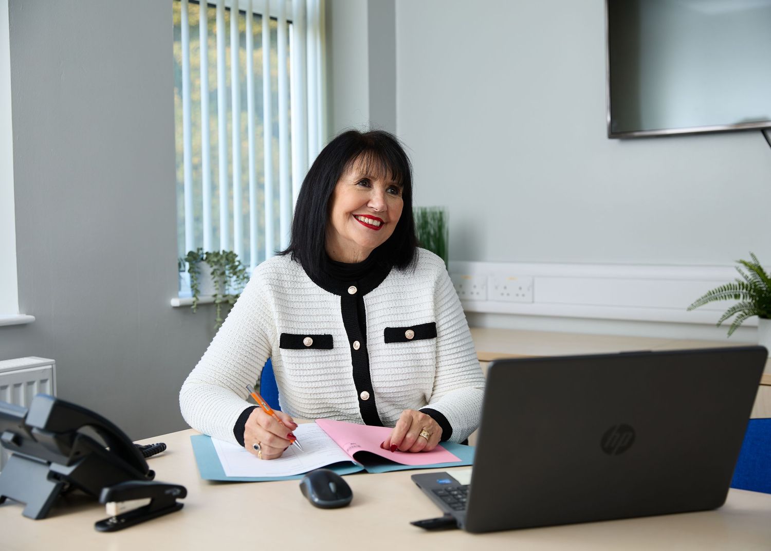 Professional woman in white blazer smiling at desk with HP laptop and pink notepad in modern office.