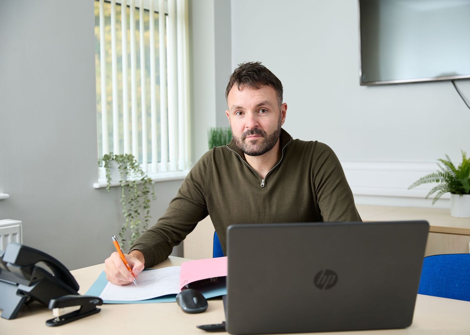 Focused bearded man in olive sweater working at desk with HP laptop and documents in modern office.
