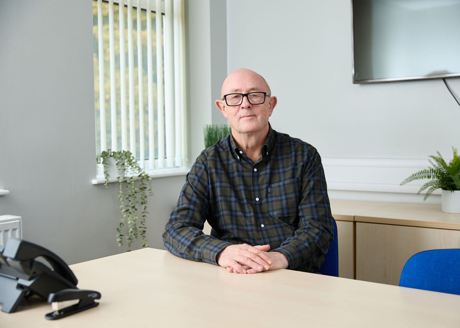 Bespectacled senior businessman sitting at wooden desk in bright modern office environment.