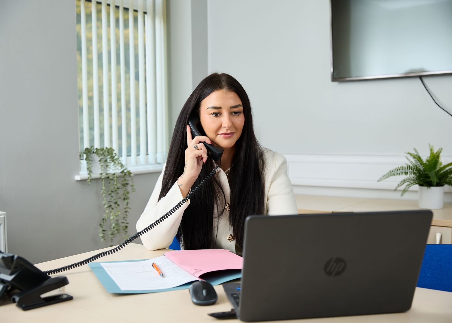 Professional woman talking on phone at office desk with HP laptop, folders, and plant in bright modern workspace.