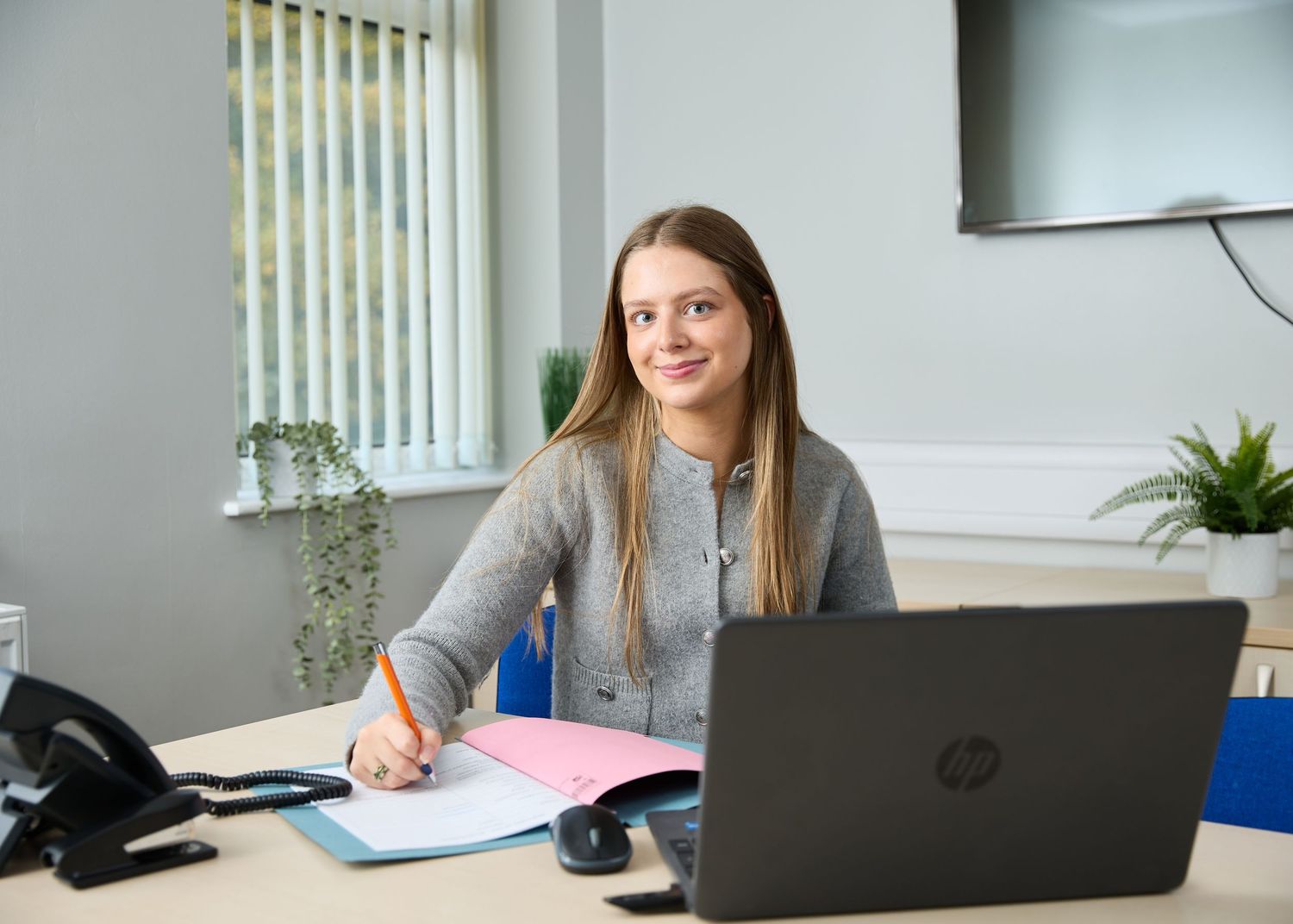 Young woman smiling at desk with HP laptop and notebook in bright office.