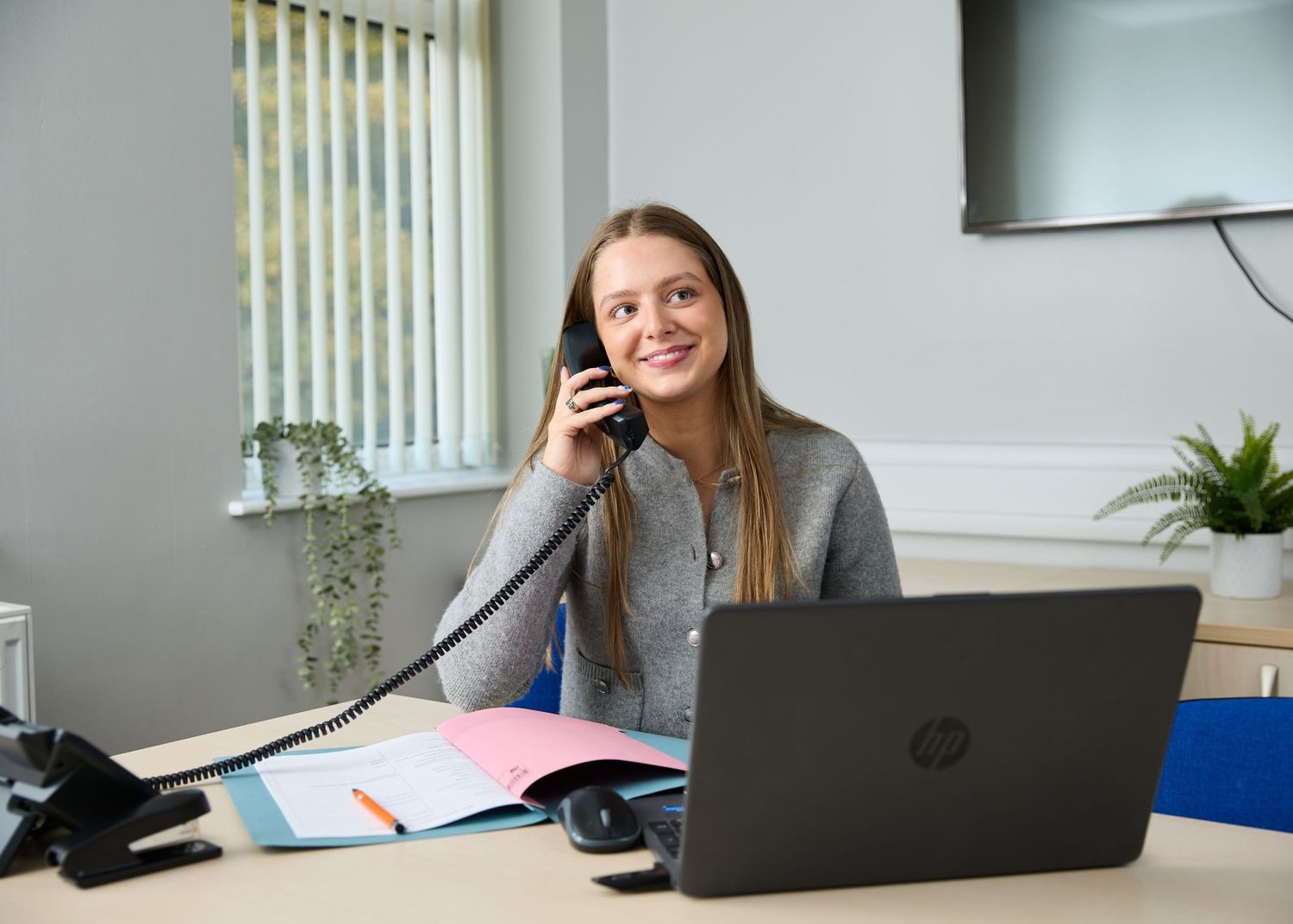 Young woman smiling while talking on office phone, sitting at desk with HP laptop and colorful folders.