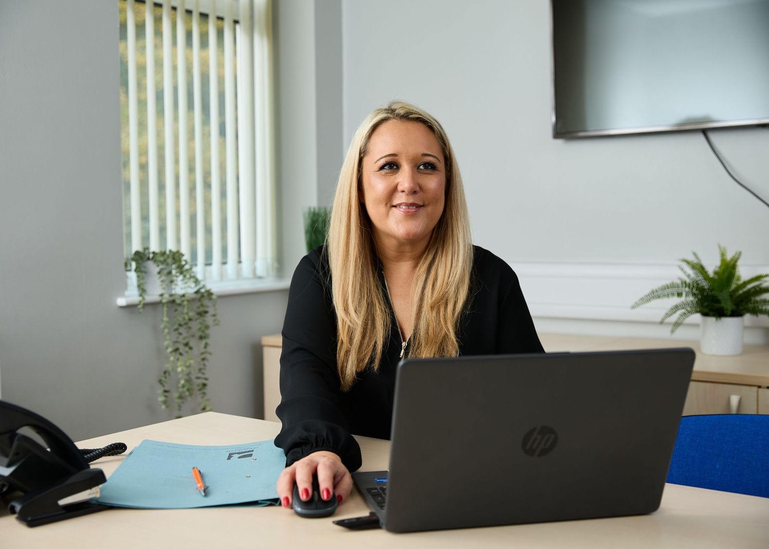 Professional woman with blonde hair sitting at office desk with HP laptop, smiling in modern workspace.
