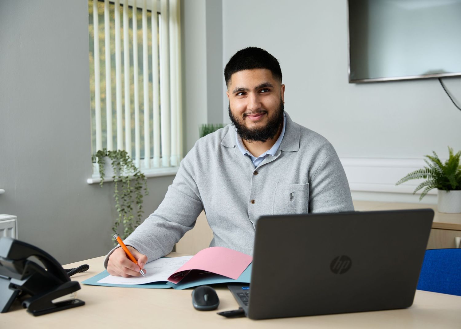 Professional man sitting at office desk with HP laptop, writing in notebook, wearing grey shirt.