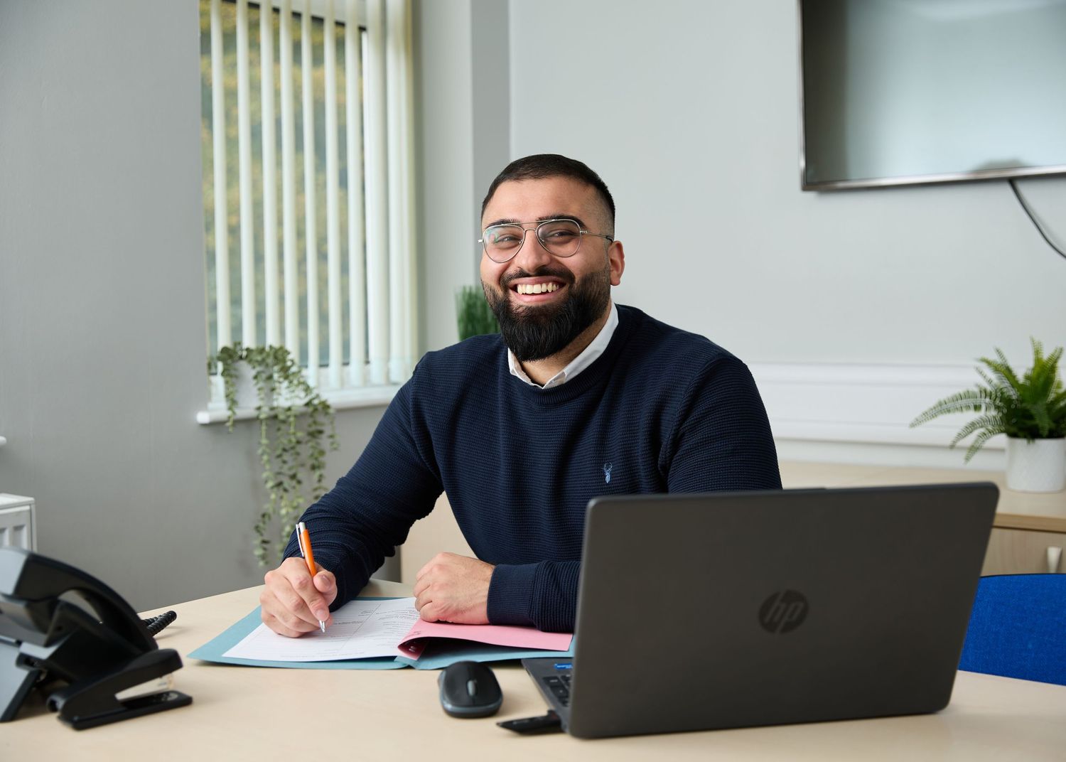 Smiling bearded man in navy sweater sitting at desk with HP laptop and documents in bright office.