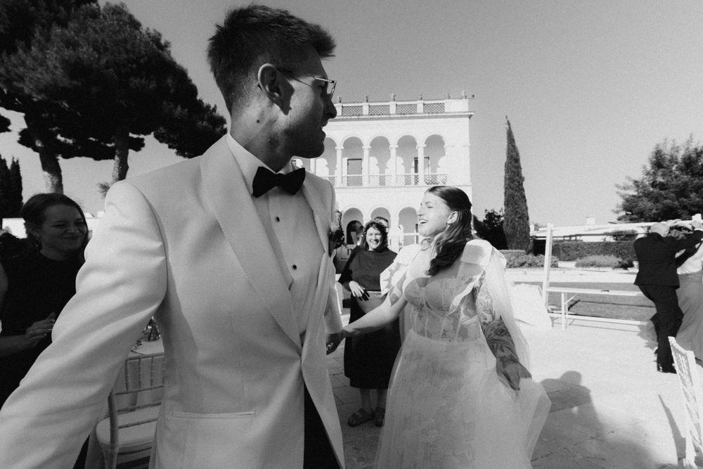 Black and white photo of a groom in white tuxedo dancing with bride in elegant dress at outdoor wedding venue.