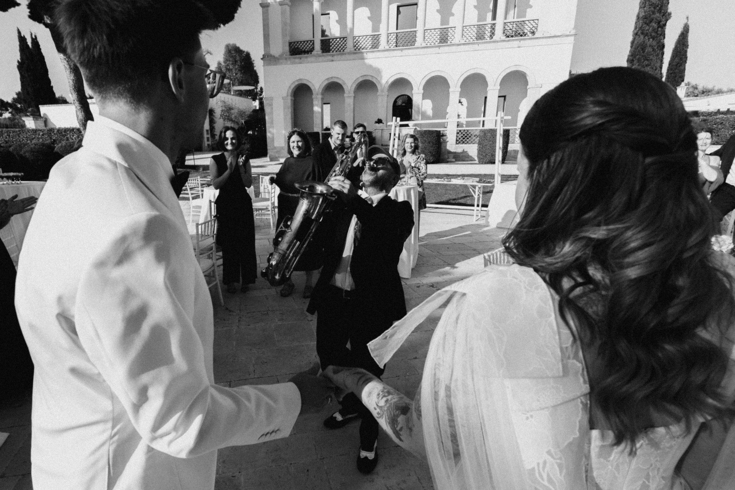 Black and white photo of wedding guests celebrating with a brass band outside a grand villa.