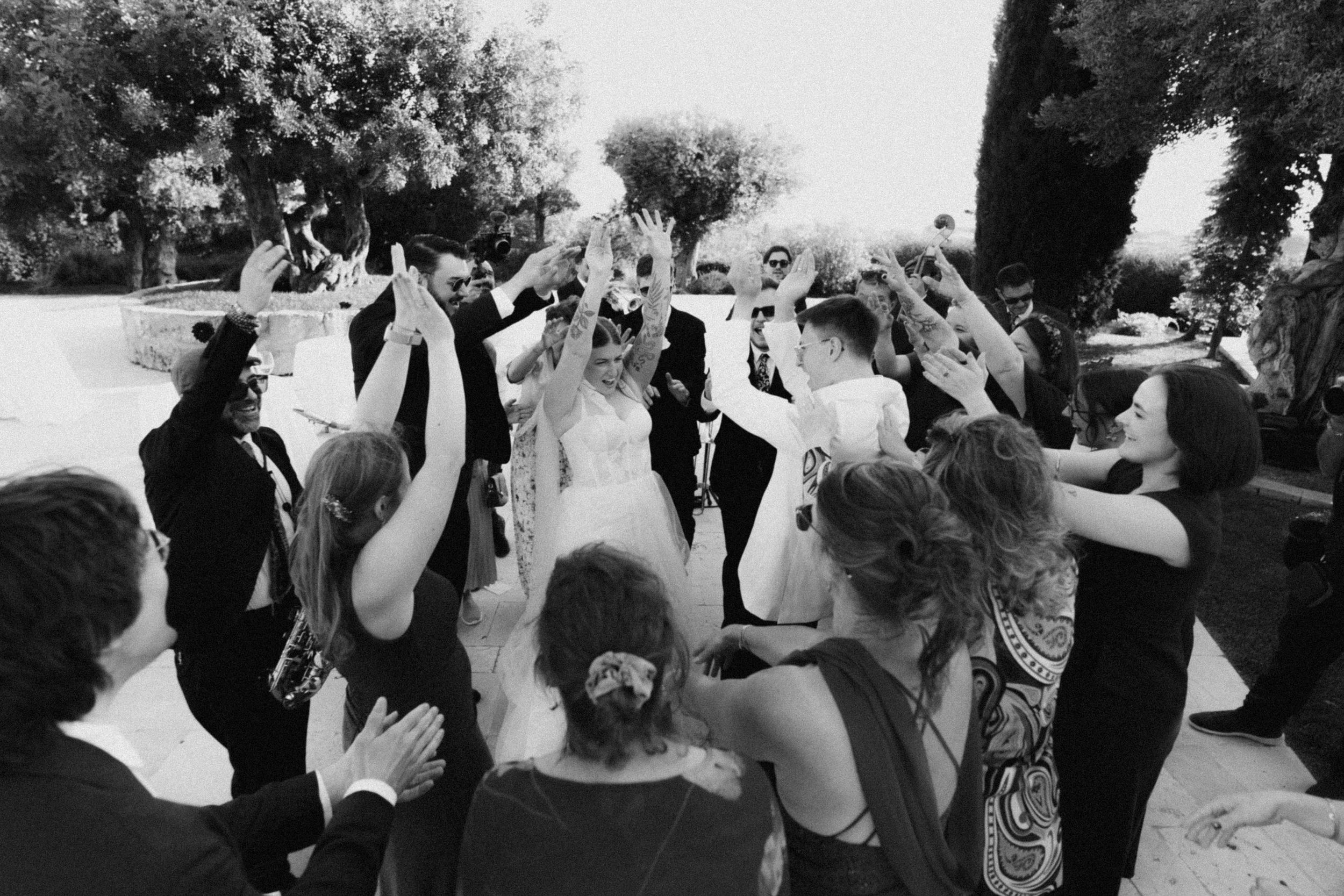 Black and white wedding photo of bride and groom celebrated by guests raising hands in joyful outdoor ceremony.