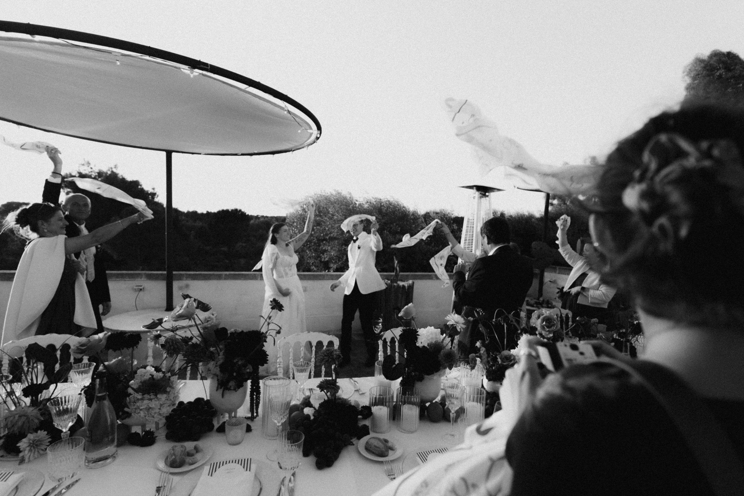 Black and white photo of guests celebrating at an outdoor wedding reception table with white napkins waving.