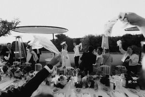 Black and white photo of wedding guests raising napkins at an elegant outdoor reception dinner table celebration.