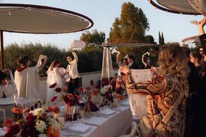 Outdoor wedding reception with guests raising napkins, vibrant red and orange floral centerpieces on a long table.