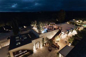 Aerial night view of Mastroni venue with illuminated terrace, lush trees, and elegant outdoor dining area under dark sky.