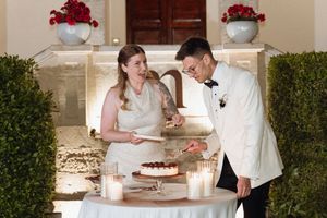 Bride and groom cutting a chocolate wedding cake at candlelit table outdoors, surrounded by lush hedges and red floral arrangements.