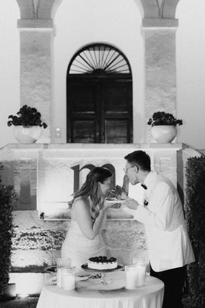 Black and white photo of a bride and groom sharing wedding cake outdoors at an elegant venue at night.