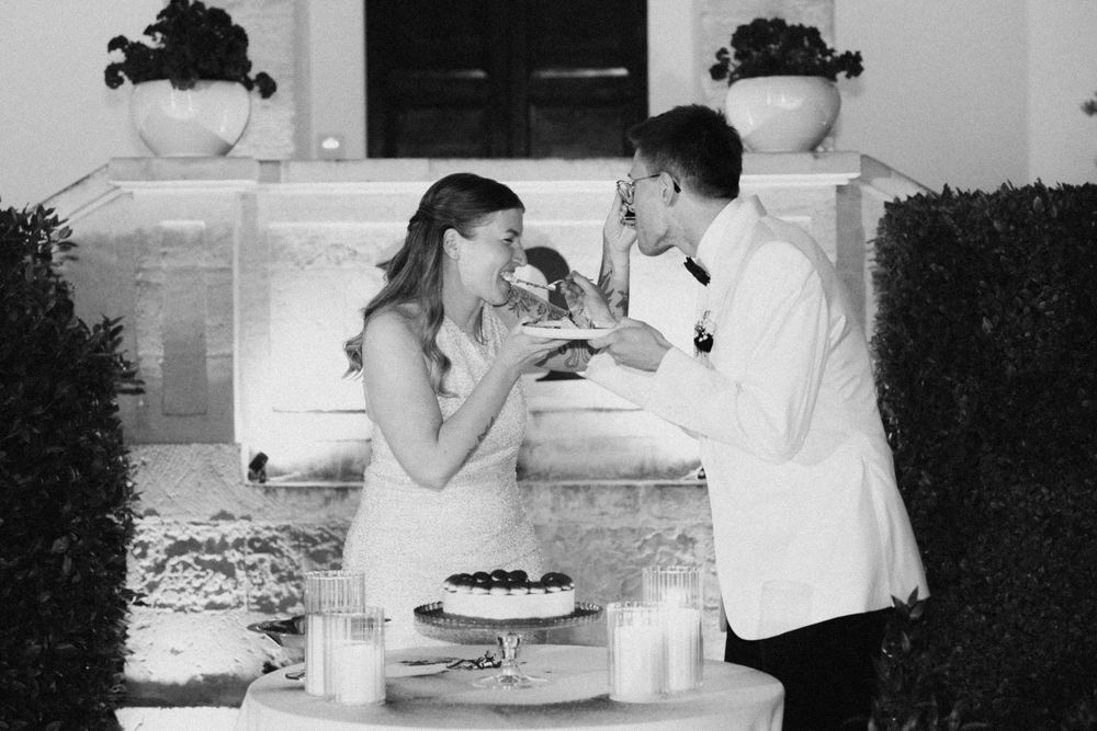 Black and white photo of a bride and groom sharing wedding cake at a candlelit outdoor reception table.