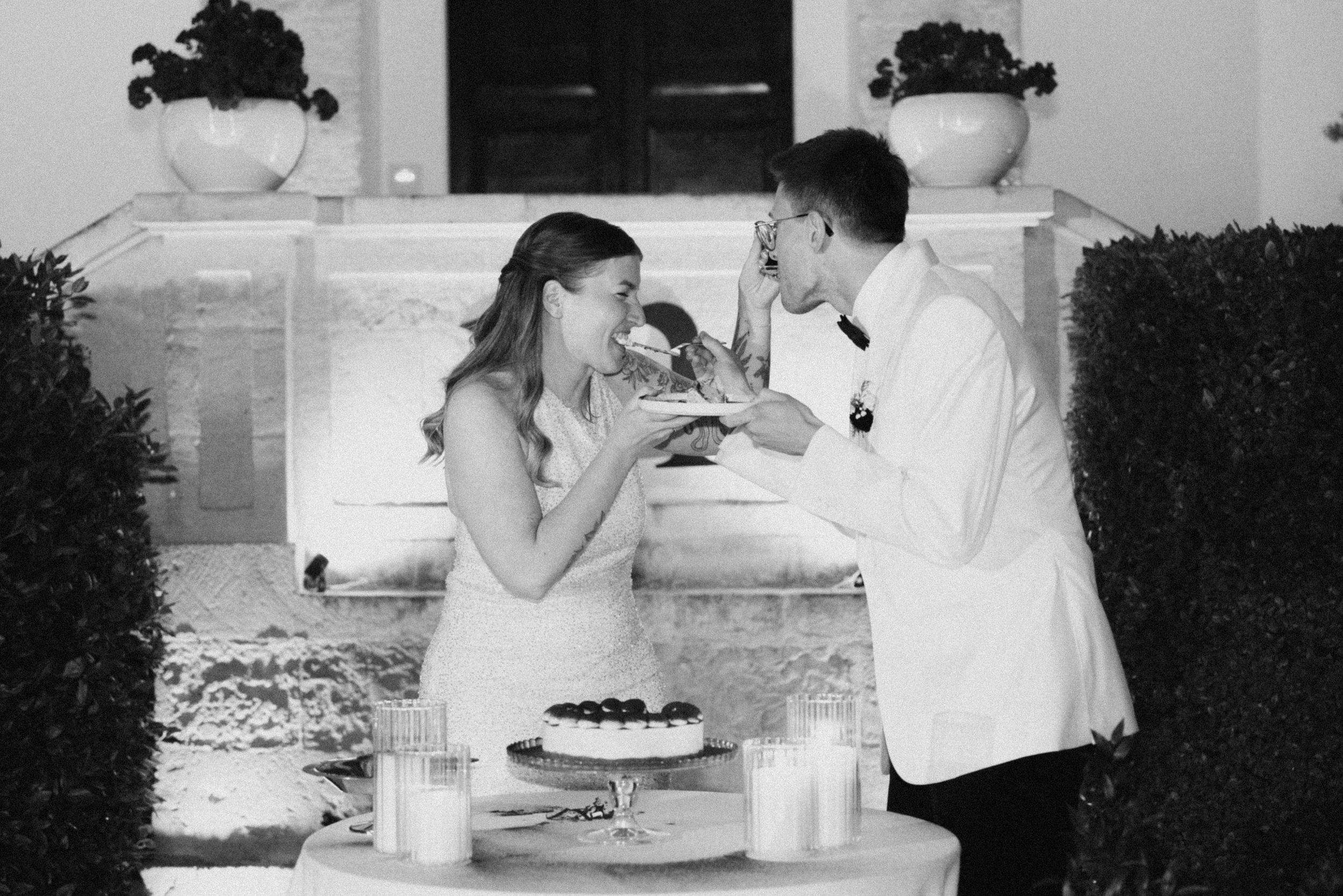 Black and white photo of a bride and groom sharing wedding cake at a candlelit outdoor reception table.