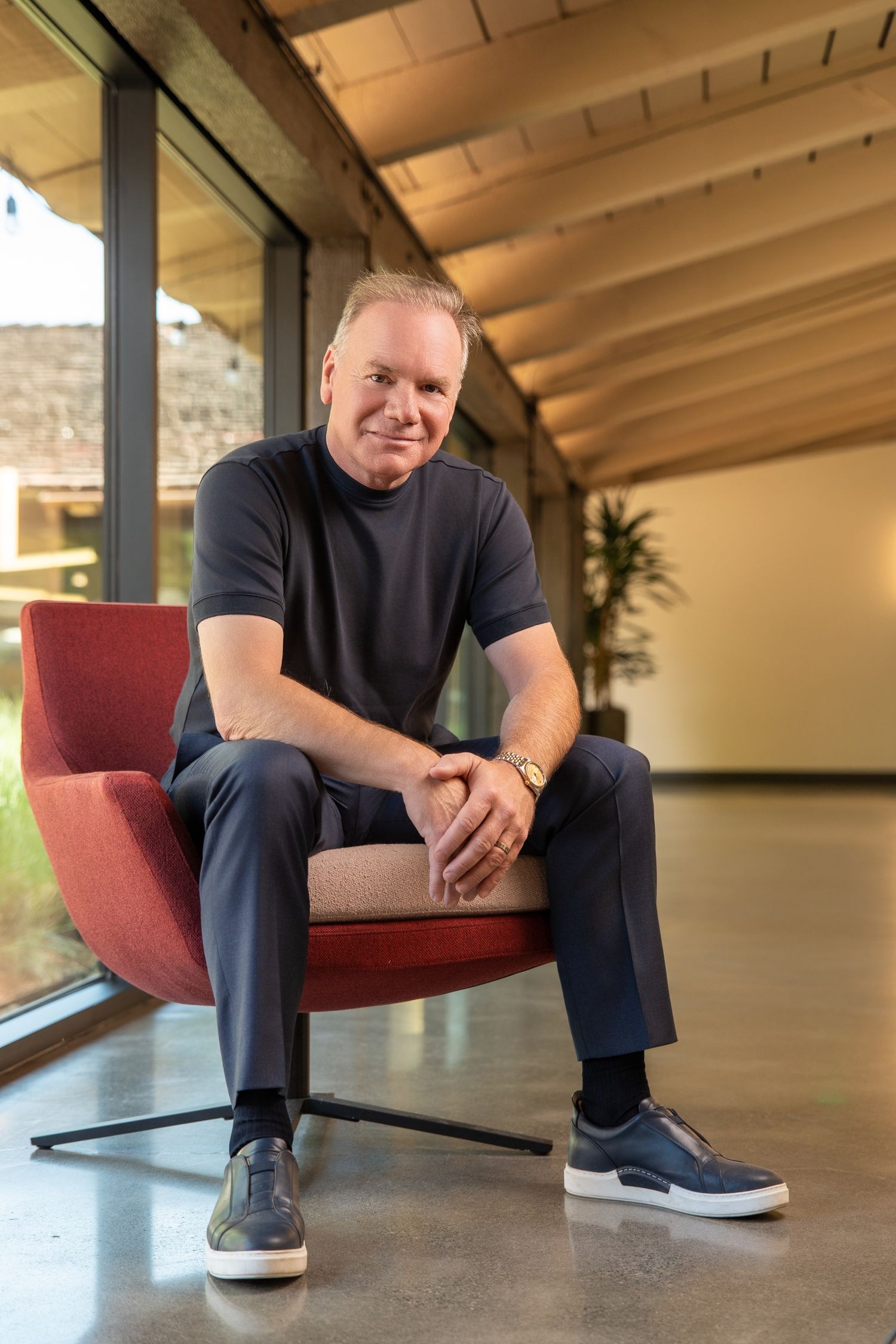 Middle-aged man in dark casual outfit sits smiling in red chair inside modern building with wood ceiling.