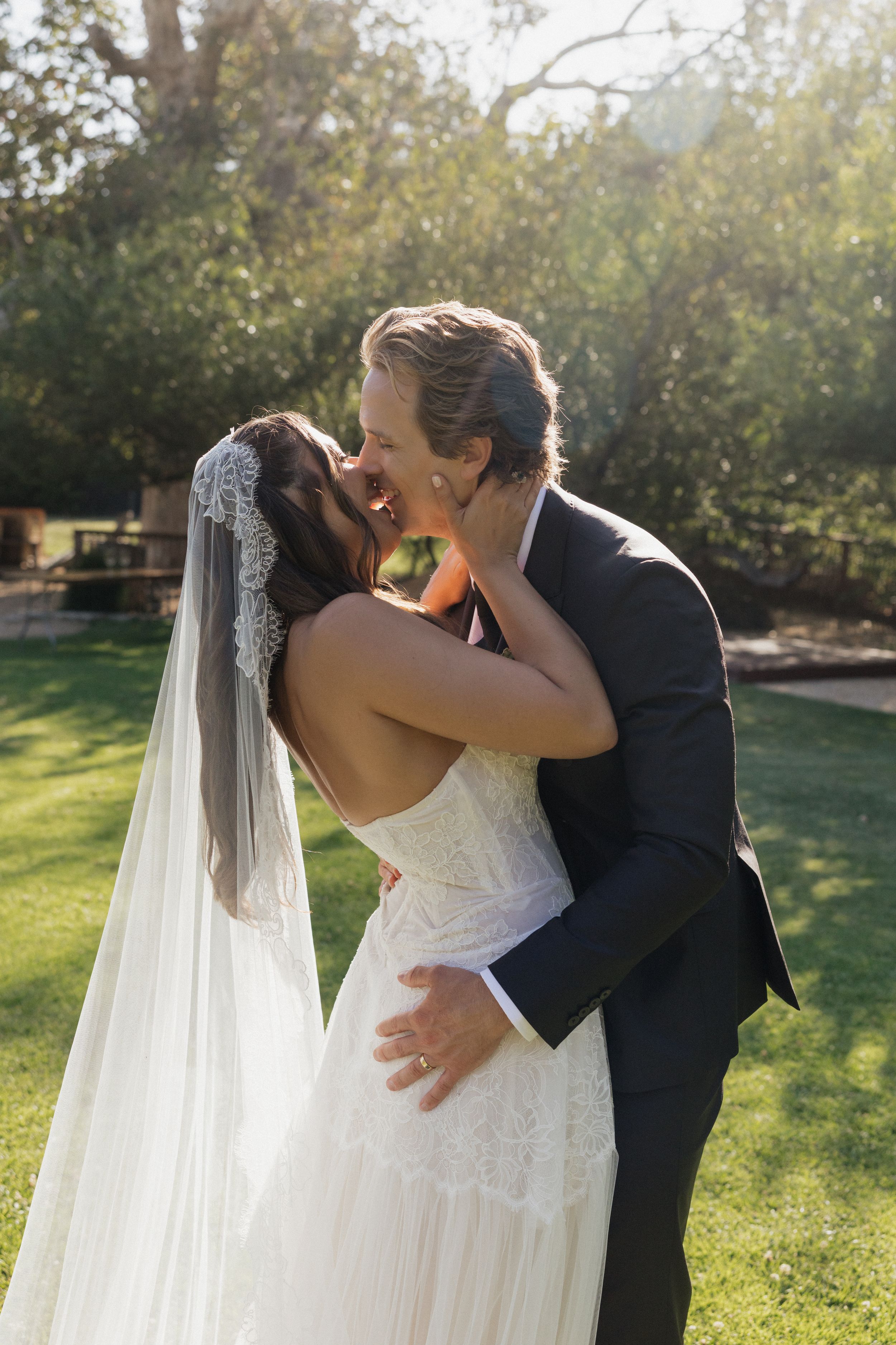 Bride and groom sharing a romantic kiss outdoors on green lawn, bride wearing lace wedding dress and veil.