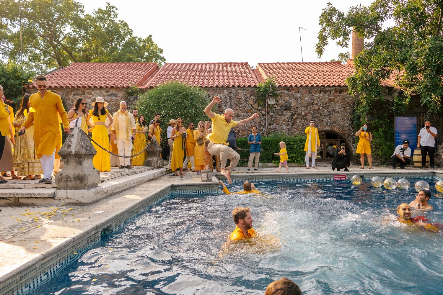 People in yellow outfits celebrate at a pool party, with one person jumping in while others swim and watch.