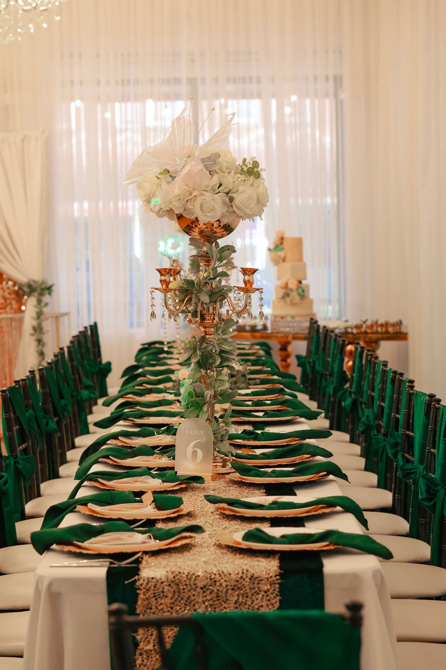 Elegant wedding reception table with emerald green napkins, gold chargers, sequin runner, and tall floral centerpiece.