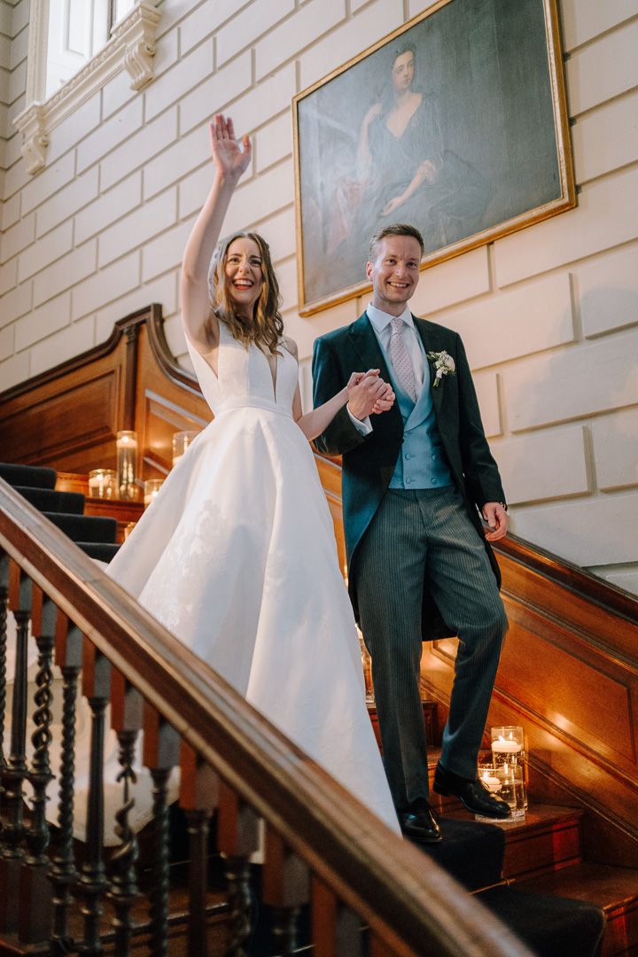 A joyful bride in a white gown waves while holding hands with her groom in a dark suit on an elegant staircase.