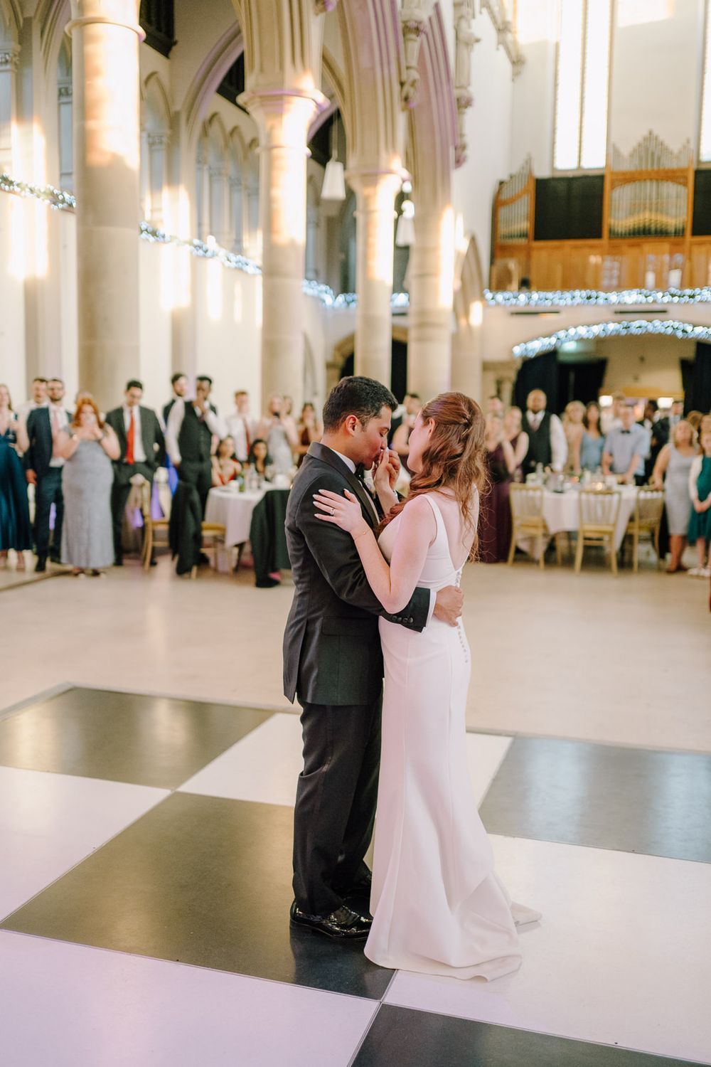 Bride and groom share first dance on checkered floor in elegant church wedding reception with guests watching.