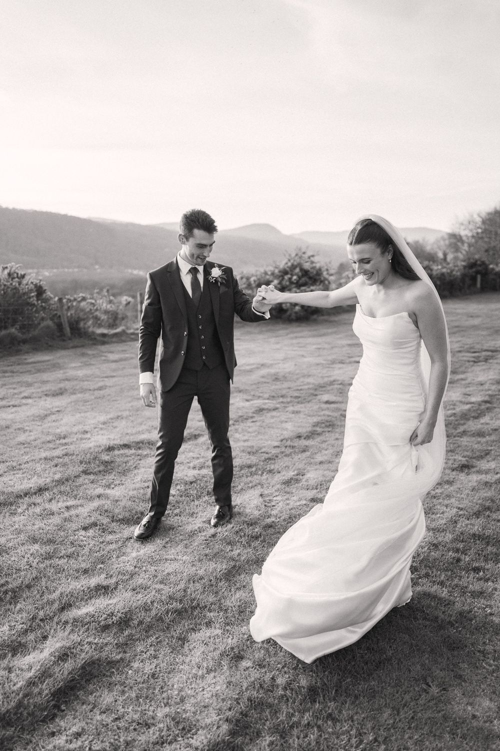 Black and white photo of bride and groom holding hands walking on grass with mountain backdrop.