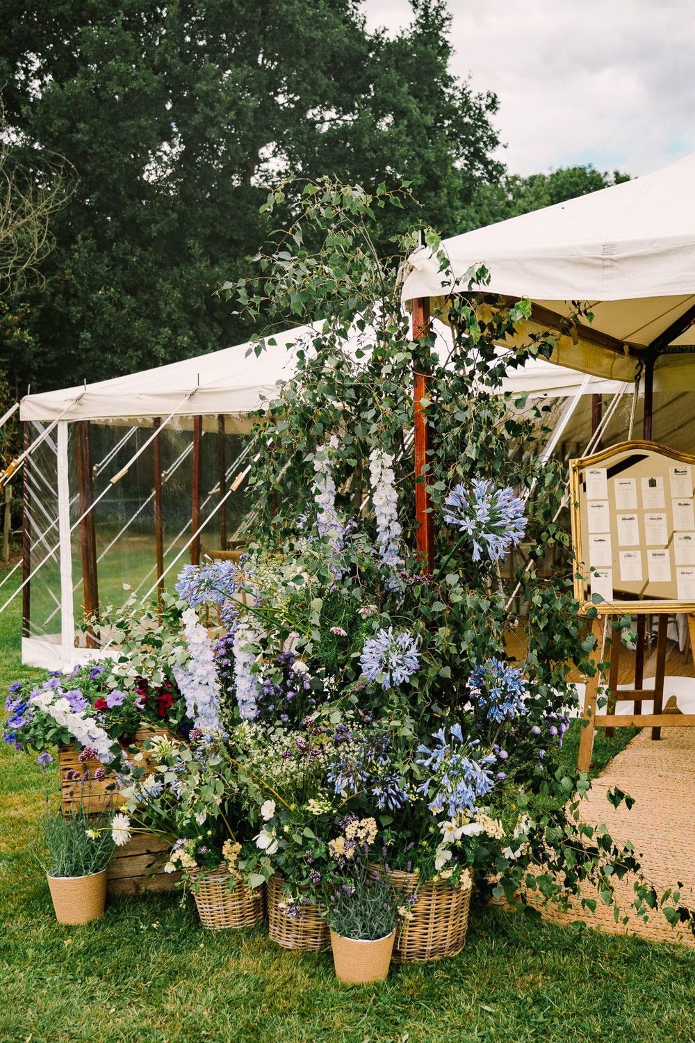 Elegant outdoor wedding floral display with blue delphiniums and greenery in wicker baskets beside a marquee tent.
