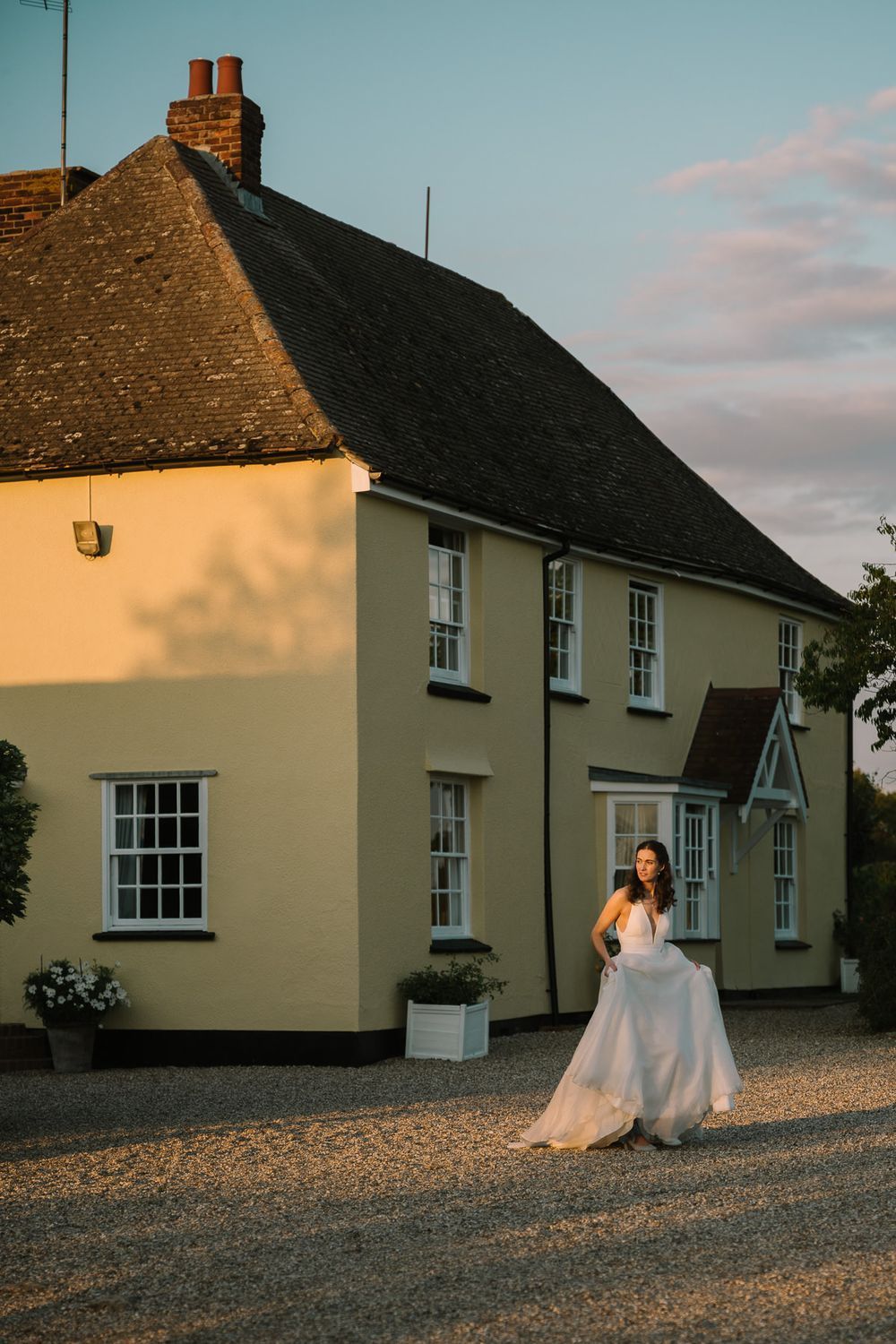 Bride in flowing white gown stands on gravel drive before golden-lit yellow country house at sunset.