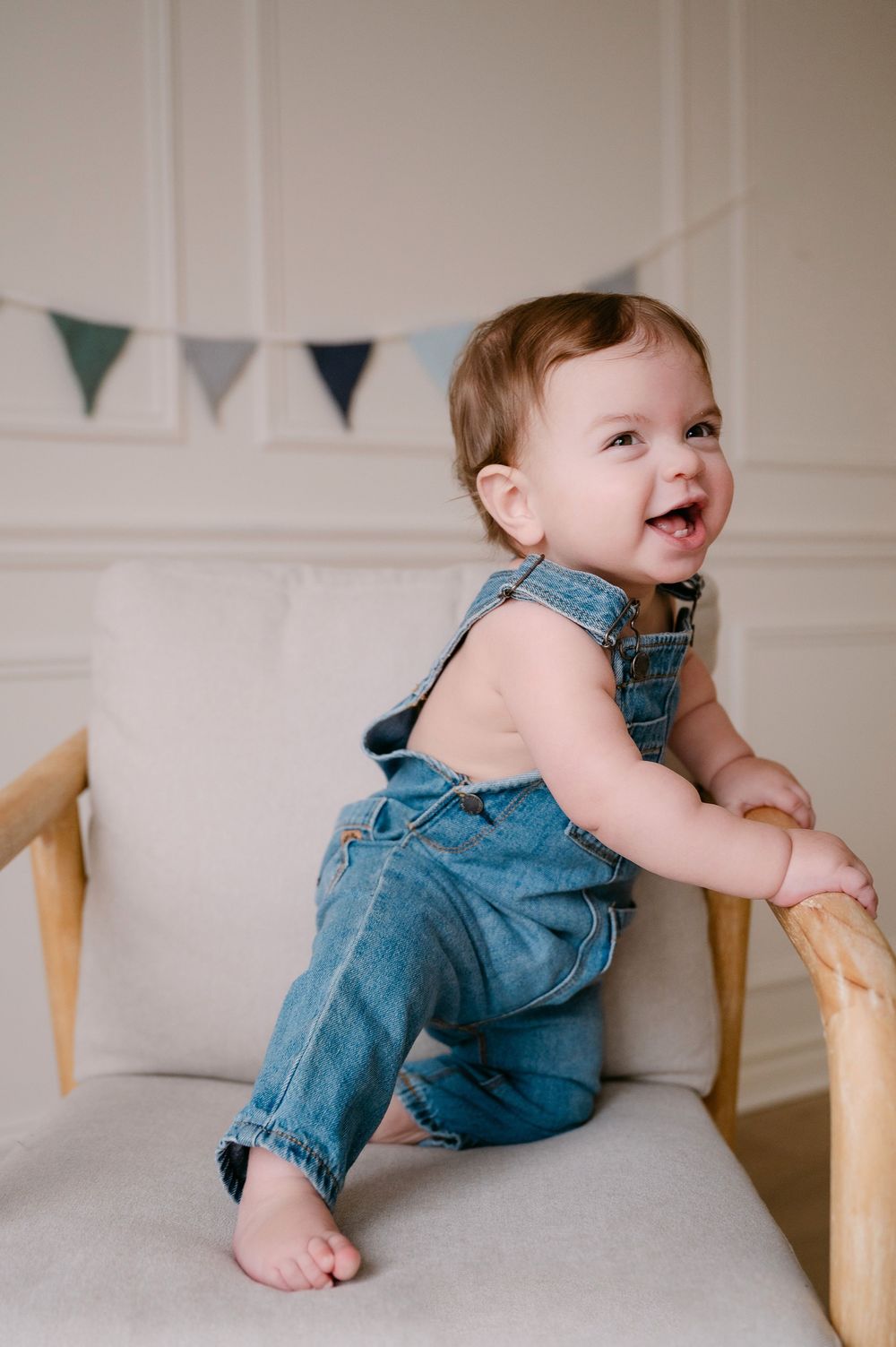 A happy baby in denim overalls stands on a white chair, smiling joyfully with bunting flags in the background.