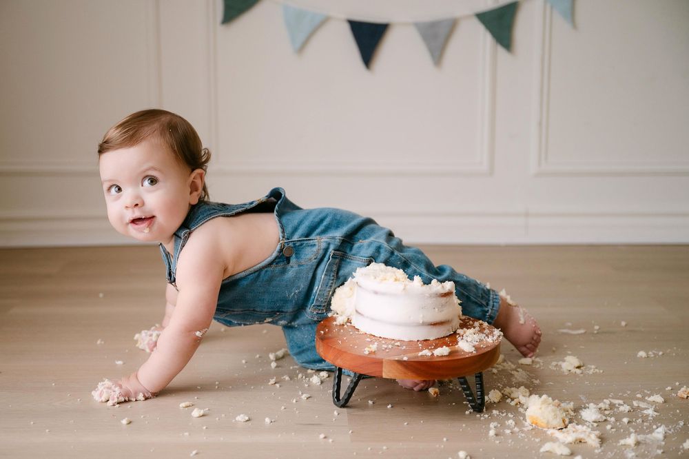 Baby in denim overalls crawling near a smashed first birthday cake on a wooden stand during a cake smash photo session.