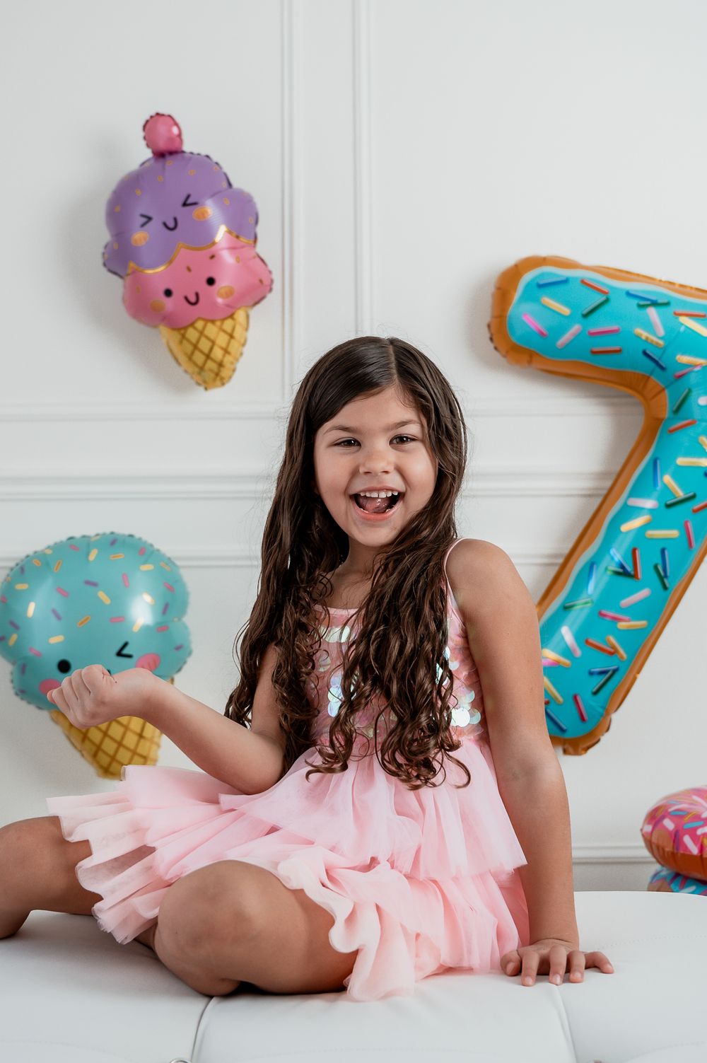 Happy girl in pink dress sitting with ice cream and number 7 balloons at a birthday party celebration.