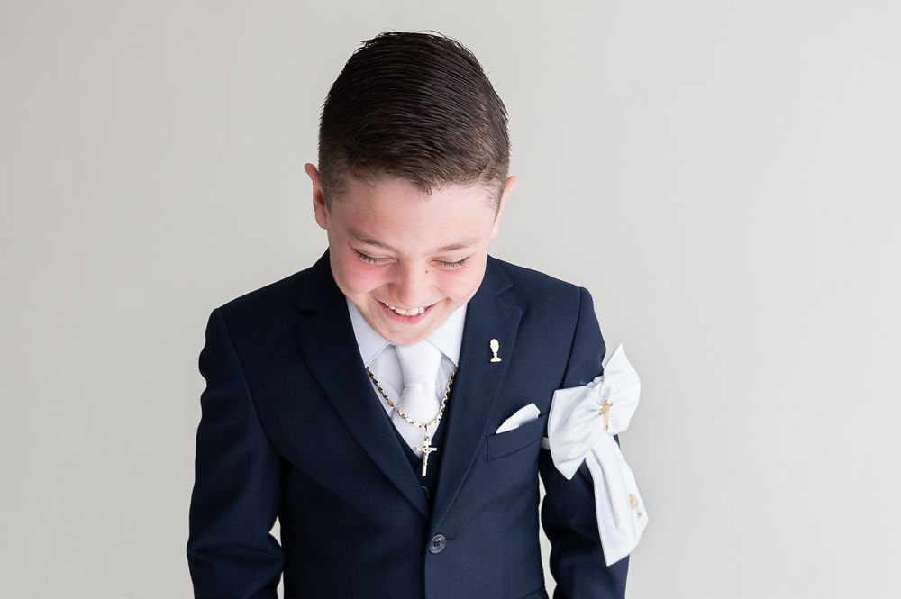 A smiling boy in a navy blue suit with white pocket square and tie, dressed for First Communion.