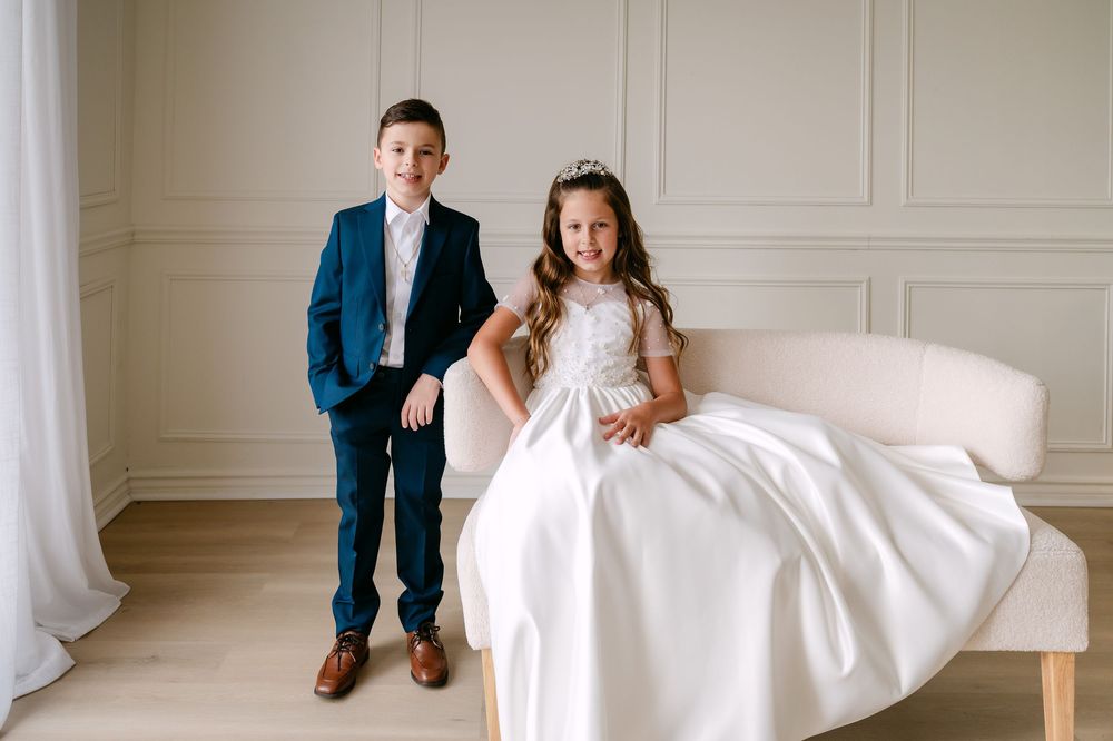 Boy in blue suit and girl in white dress with crown sit together in elegant white room for formal portrait.