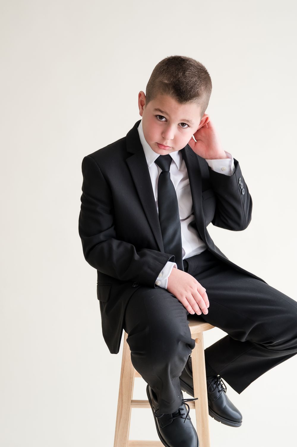 Young boy in black suit and tie sitting on wooden stool against white background, posing formally.
