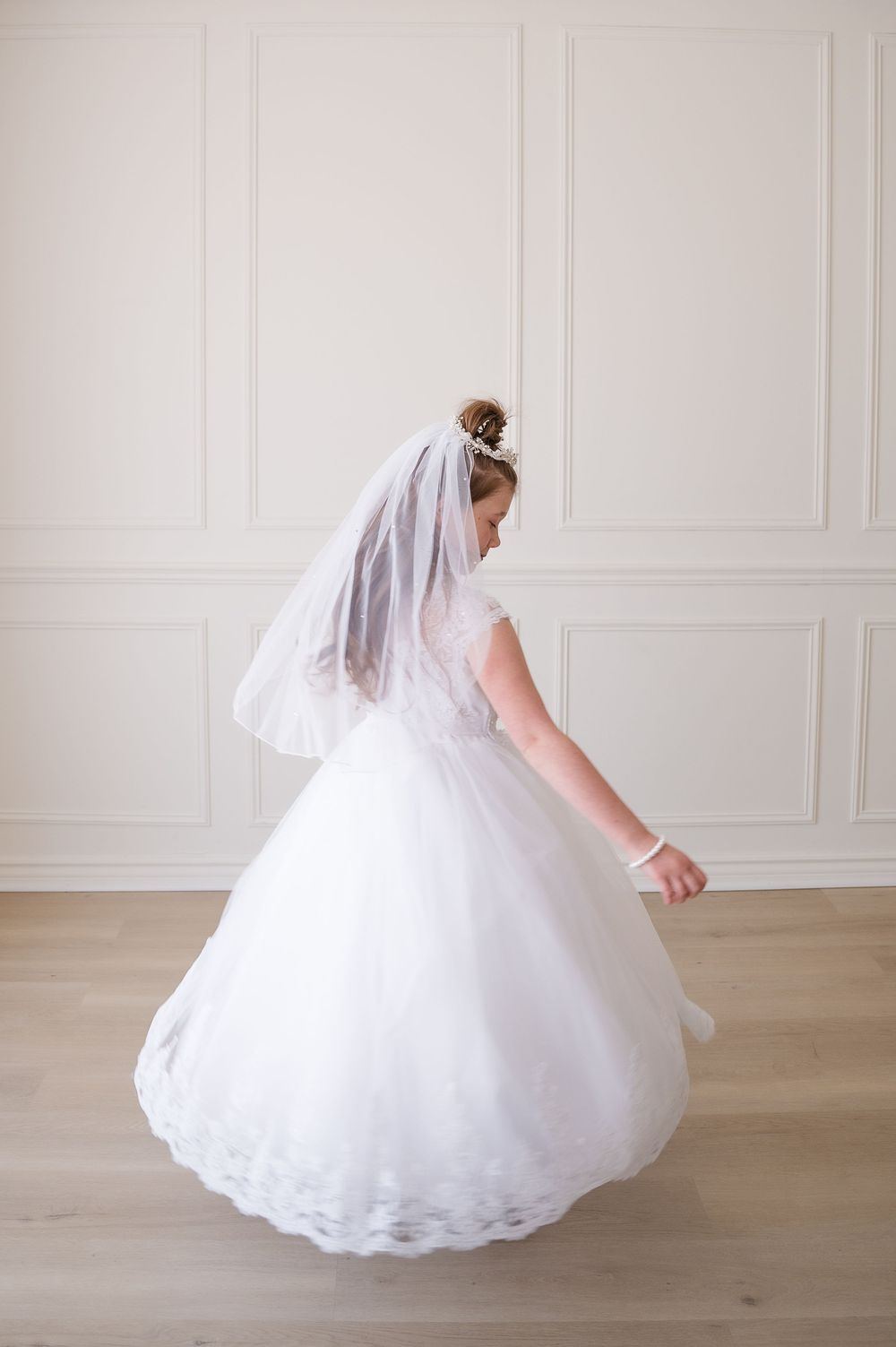 Young girl in white First Communion dress and veil twirling in bright room.
