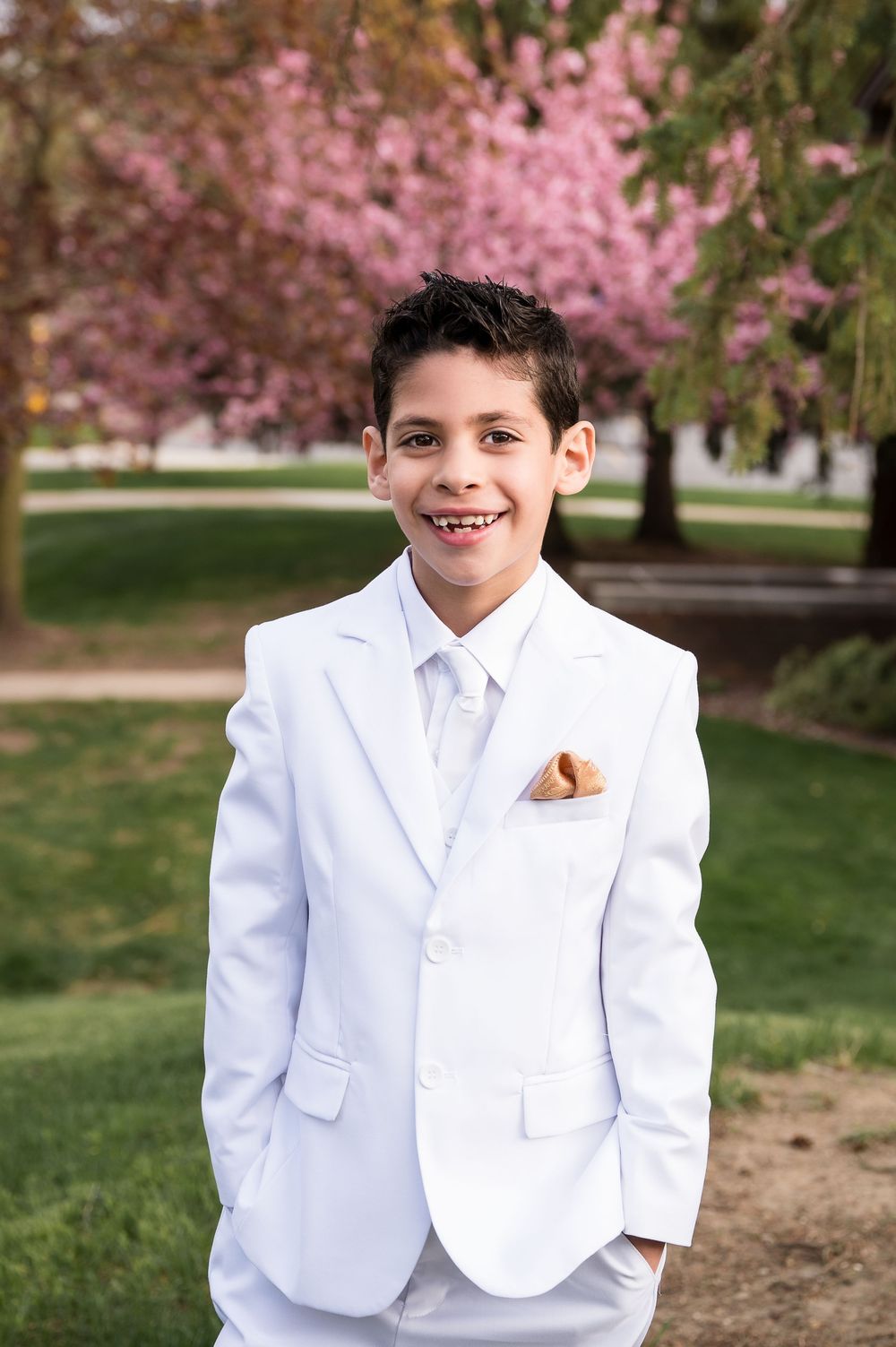 A smiling boy in a white suit with tie and pocket square poses outdoors near blooming pink cherry blossom trees.