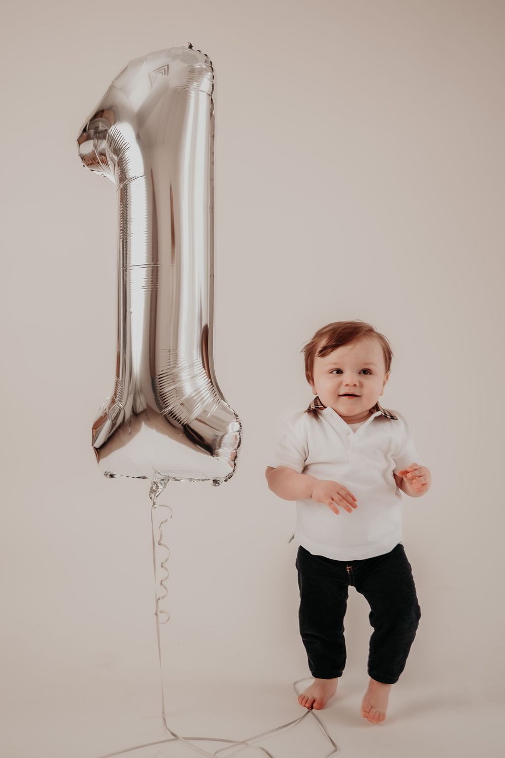 A smiling baby in white shirt and dark pants stands next to a large silver number 1 balloon for their first birthday.