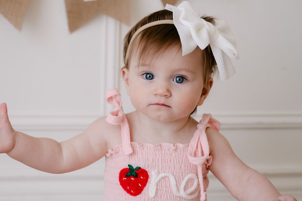 Adorable baby girl in pink smocked strawberry romper with white bow headband celebrating her first birthday.
