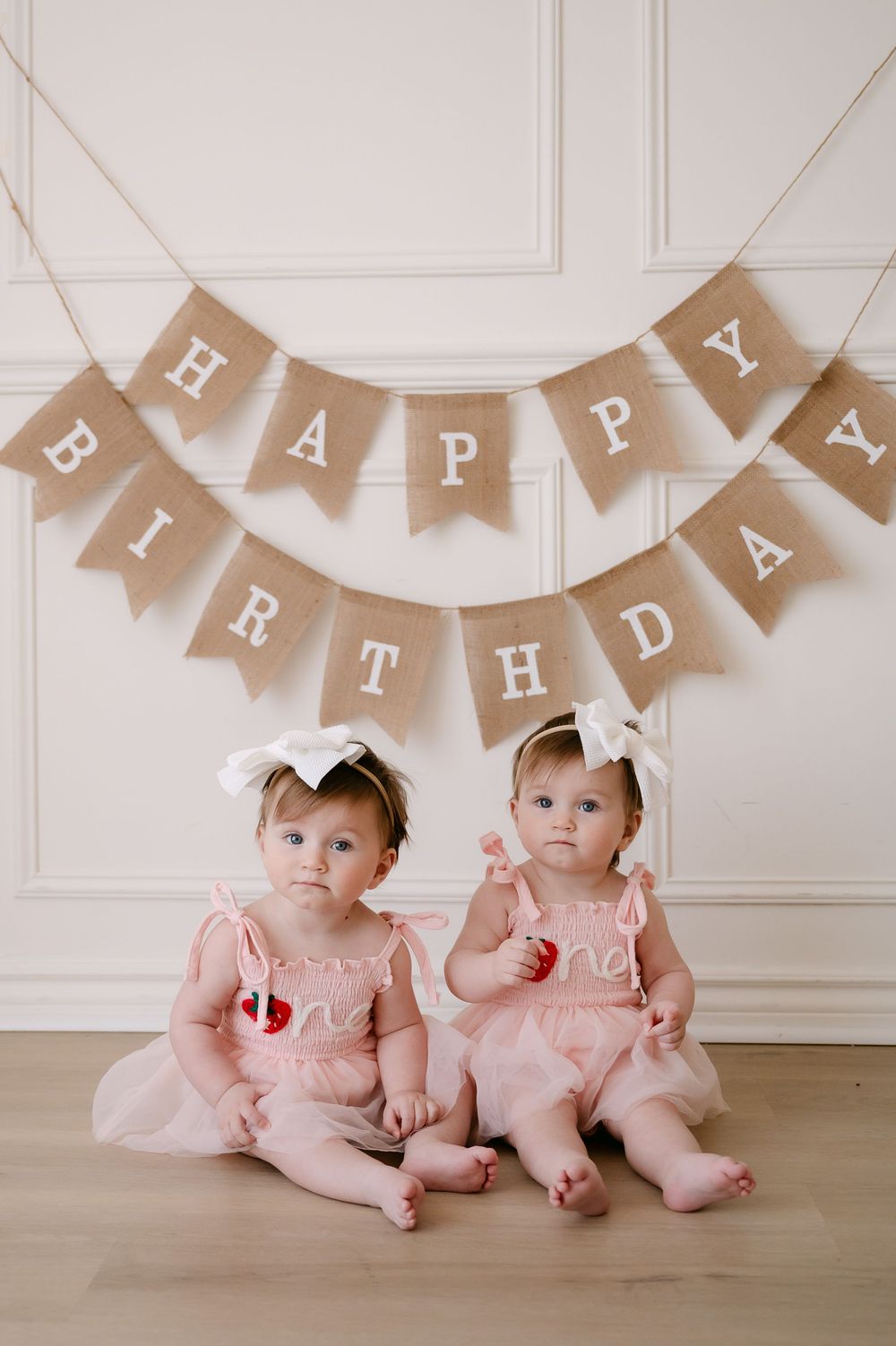 Twin baby girls in pink dresses with white bows sit before a burlap Happy Birthday banner for their first birthday.