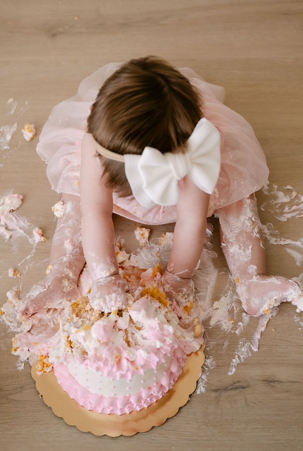 Toddler girl in pink tutu smashing first birthday cake on wooden floor, cake smash photography.