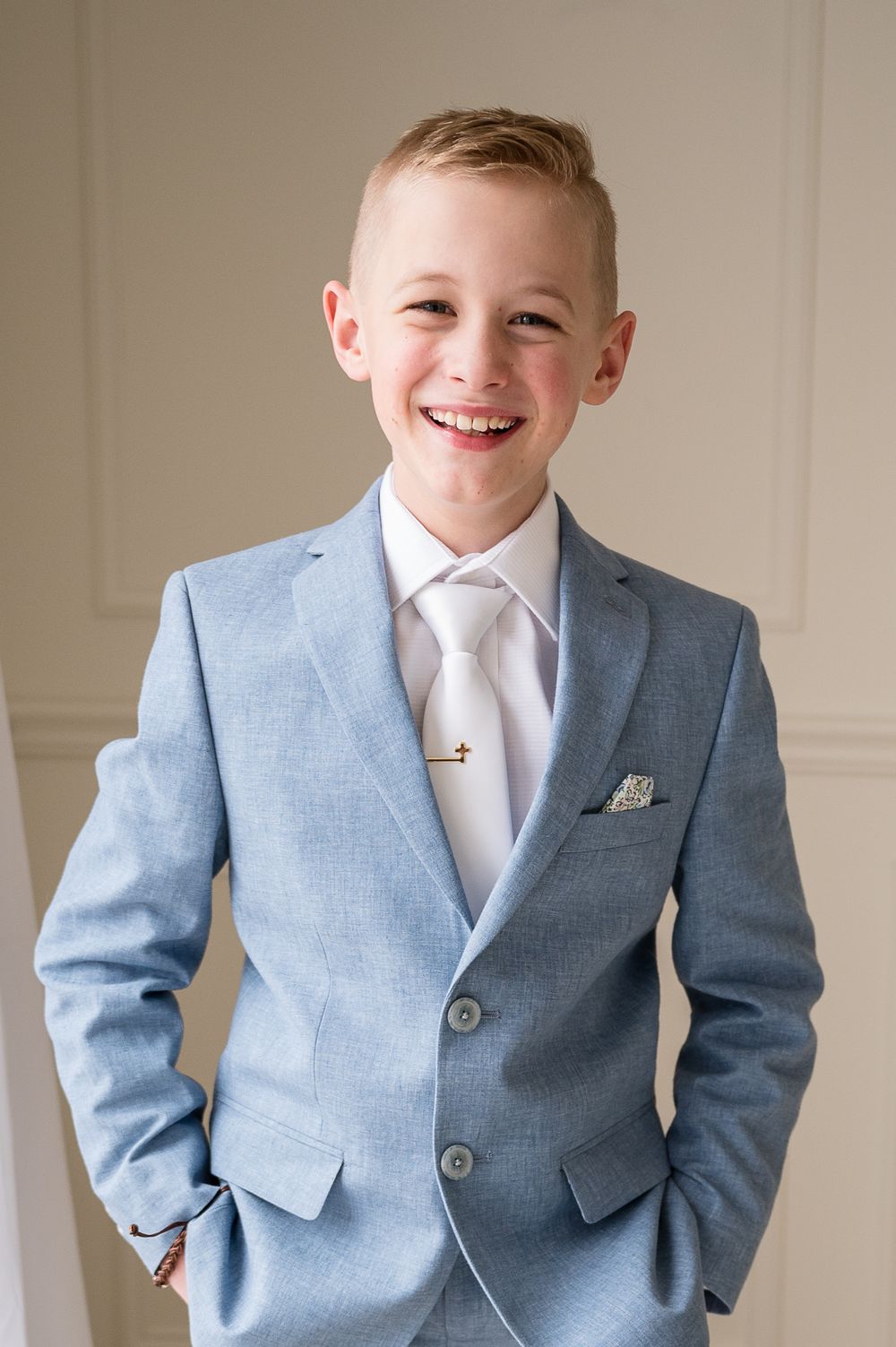 Smiling boy in a light blue suit with white tie and pocket square, dressed formally for a special occasion.