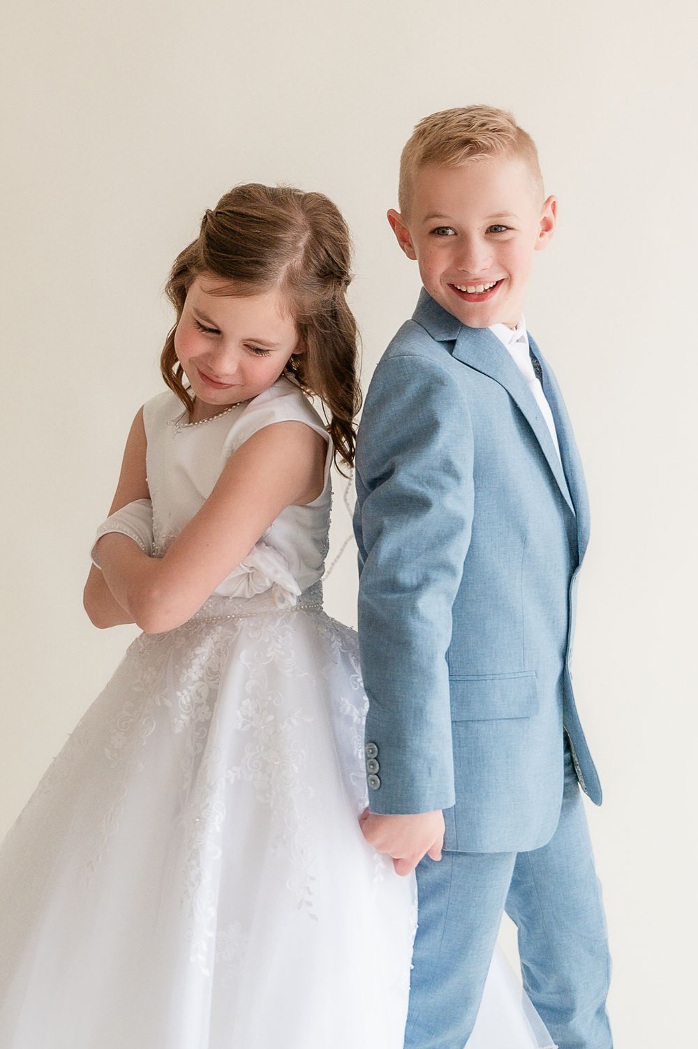 Two children in formal wear, a girl in white dress and boy in blue suit, pose back-to-back for a portrait photo.