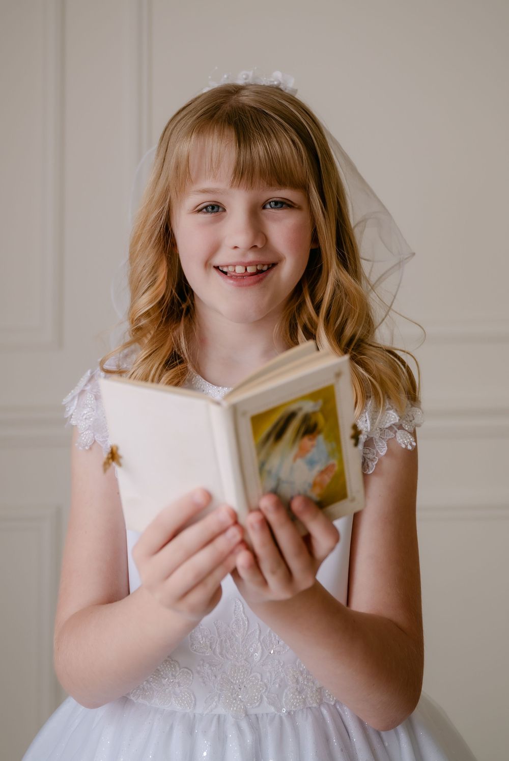Young girl in white lace dress and veil holds a prayer book, smiling during her First Holy Communion.