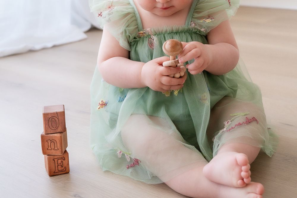 Baby in green floral dress sitting on floor playing with wooden alphabet blocks and small toy.