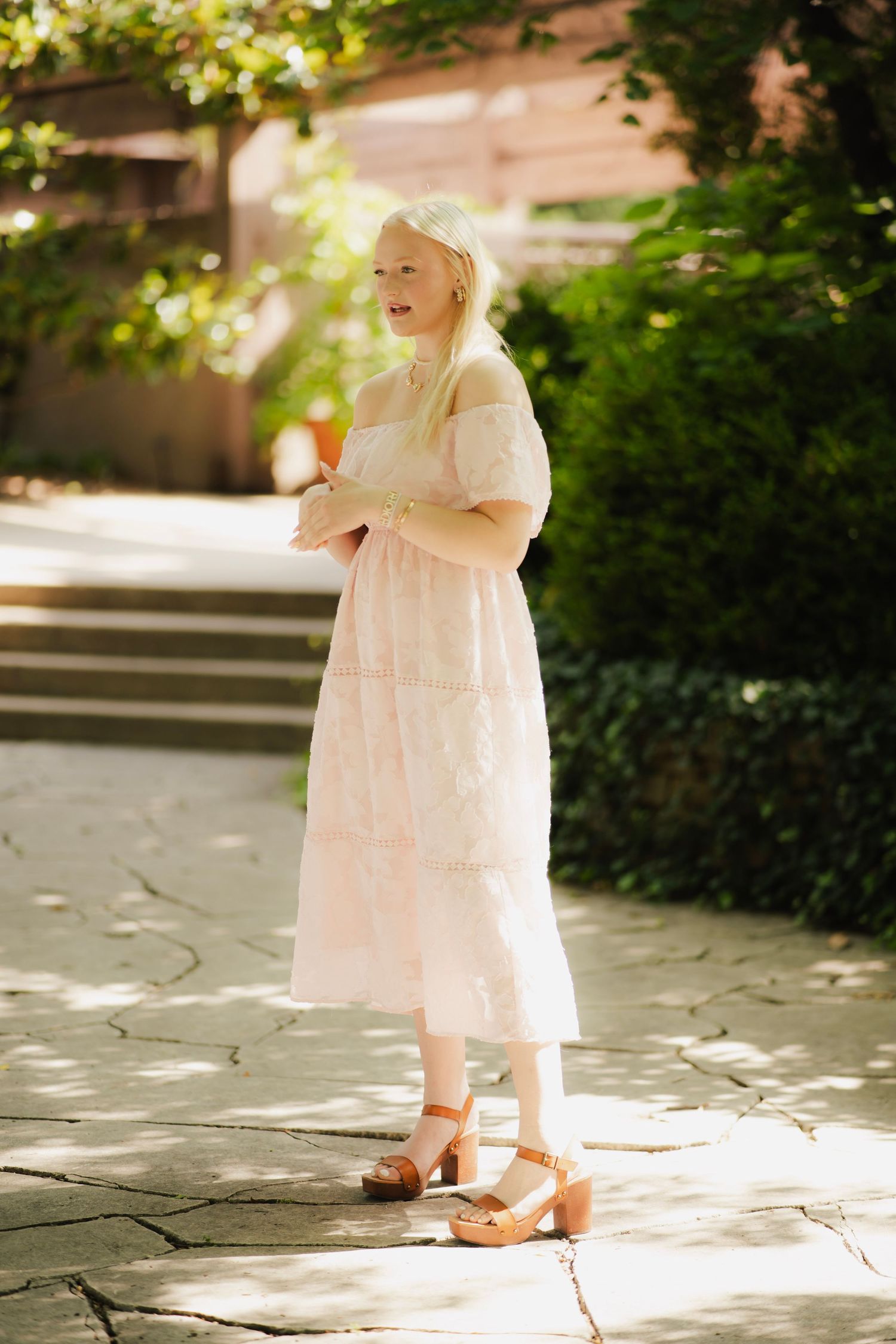 Young blonde woman in white lace midi dress with block heels standing on stone path in garden.