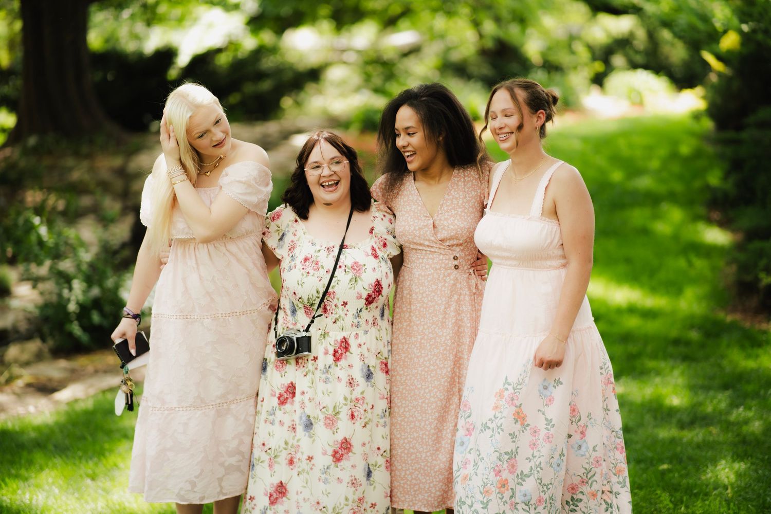 Four smiling women in pastel floral dresses embracing outdoors in a bright green garden setting.